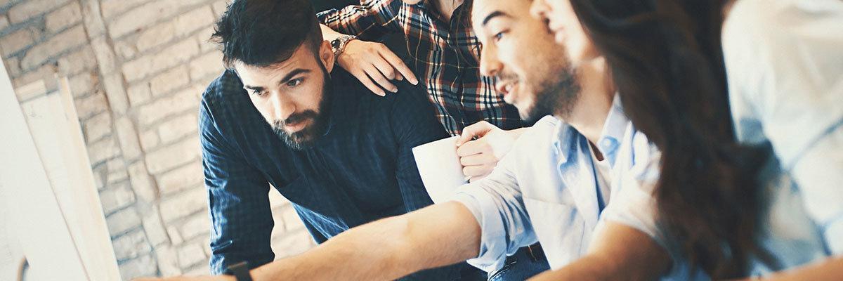 Closeup side view of group of IT experts completing a task in their office. There are two men and two women gathered around dual screen computer. One of the men is leading the project.