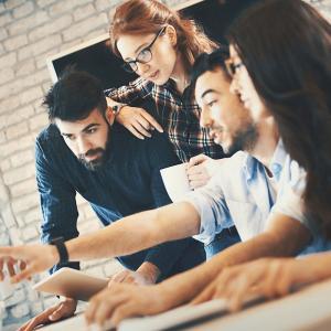 Closeup side view of group of IT experts completing a task in their office. There are two men and two women gathered around dual screen computer. One of the men is leading the project.